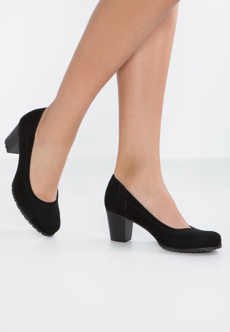 Jambes portant des chaussures en daim noir à talon bas bloc sur fond blanc, montrant un pied levé, l’autre à plat sur le sol.