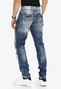 Blaue verwaschene Jeans mit einzigartigen gemusterten Gesäßtaschen, die ausgefranste Kanten und eine faded Waschung aufweisen. Getragen mit weißen Turnschuhen.