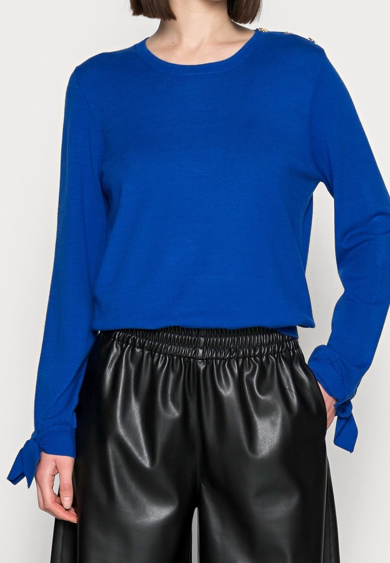 Femme portant un haut bleu vif à manches longues avec des boutons aux épaules et des poignets noués, associé à un pantalon noir taille haute en similicuir.