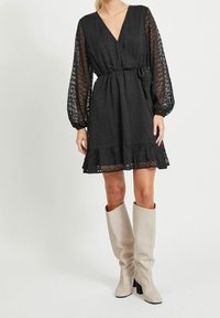 Svart omlottklänning med långa, genomskinliga spetsärmar, volangkant och tygbälte. Kombineras med höga, off-white läderstövlar med kraftiga klackar.