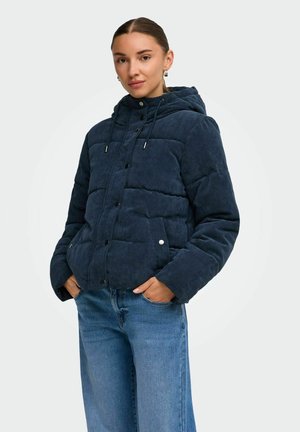 Femme portant une veste matelassée en velours côtelé bleu marine avec capuche et un jean bleu, debout les mains dans les poches, fond neutre.