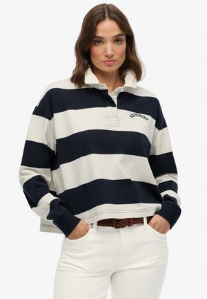 Navy and cream striped rugby-style shirt with a collar, featuring an embroidered logo and long sleeves, paired with white pants and a brown belt.