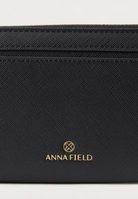 Čierna peňaženka s textúrou, vybavená zipsovým uzáverom, viacerými priehradkami na karty a zlatým logom „ANNA FIELD“.