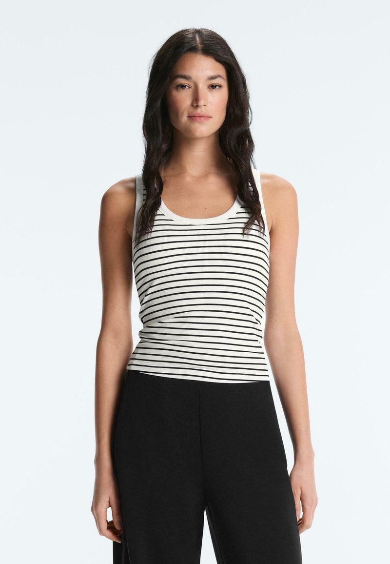 Jeune femme aux longs cheveux foncés portant un débardeur blanc à rayures horizontales noires et un pantalon noir taille haute, debout, face à l'avant.