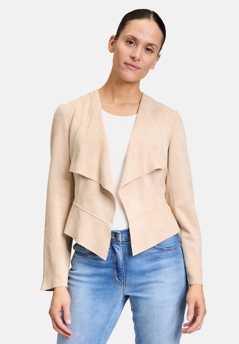 Beige Wildlederjacke mit offenem Vorderteil, geschichteten Revers, langen Ärmeln und taillierter Silhouette. Tragen über einem weißen Hemd und blauen Jeans.