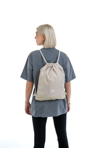 Světle béžový batoh na šňůrku s nastavitelnými popruhy, měkká látka a obdélníkový logový nášivka, nošený přes šedé tričko.