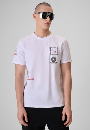 Młody mężczyzna ubrany w białą koszulkę z nadrukiem, duże czarne okulary przeciwsłoneczne z daszkiem oraz czarne spodnie, stojący na jednolitym jasnoszarym tle.