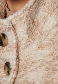 Nærbillede af beige tekstureret uldfrakke med store brune knapper og et synligt kraveområde.