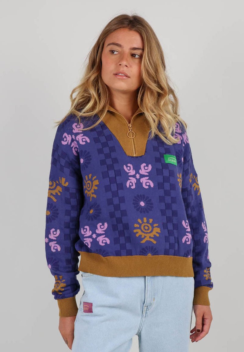 Sweatshirt violet à motifs avec un col zippé mi-hauteur, arborant des motifs géométriques en jaune et rose. Poignets et ourlet côtelés en marron.