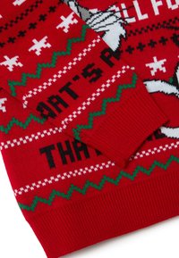 Pull en tricot rouge avec des flocons de neige blancs et des motifs zigzag verts. Le texte en noir indique "QUOI DE NEUF ?" et "C'EST ÇA."