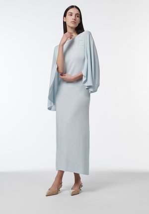 Mujer con un vestido maxi azul claro con mangas anchas y caídas sujetas por botones, calzando tacones beige de punta afilada sobre un fondo blanco.