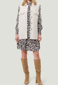 Beige ärmellose Weste mit zwei vorderen Taschen und großen Knöpfen über einem schwarz-beigen Leopardenmuster-Kleid, kombiniert mit knielangen tanfarbigen Stiefeln.