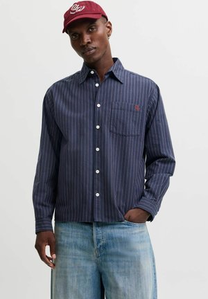 Homme portant une casquette rouge, une chemise bleu marine à fines rayures avec boutons et un jean bleu clair, une main dans la poche, sur un fond uni.