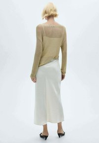 Femme blonde portant un haut beige transparent à manches longues par-dessus une robe nuisette blanche et des escarpins noirs à petit talon, debout devant un fond blanc uni.