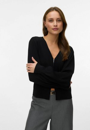 Donna con lunghi capelli castani che indossa un maglione nero con zip frontale e pantaloni grigi, in piedi con le braccia incrociate davanti a uno sfondo bianco.