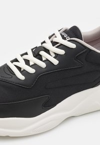 Sneaker neri con tomaia in tessuto testurizzato, lacci bianchi e suola bianca a contrasto. Gli accenti di design notevoli includono sovrapposizioni curve.
