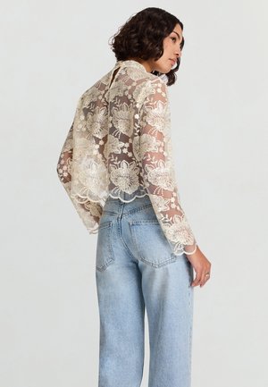 Camicia in pizzo trasparente ricamata con motivi floreali, di colore crema, orlo smerlato e maniche lunghe, abbinata a jeans in denim a vita alta azzurri.