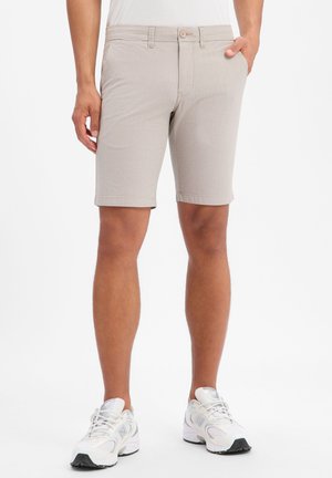 Mann trägt beige knielange Shorts und weiße Sportschuhe, steht mit einer Hand in der Tasche.