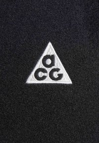 Black fabric features a white embroidered triangle with the letters "a", "c", and "g" in black. Texture appears smooth and detailed.
