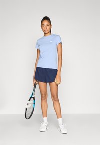 Μπλε βαμβακερό μπλουζάκι με κοντά μανίκια, συνδυασμένο με σκούρο μπλε σορτς. Το άτομο κρατά ρακέτα του τένις, φοράει λευκά αθλητικά παπούτσια και αθλητικές κάλτσες.