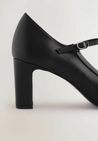 Zwarte hoge hakken met een gladde, matte afwerking. Voorzien van een enkelbandje dat wordt vastgemaakt met een kleine zilveren gesp en een stevige hak.