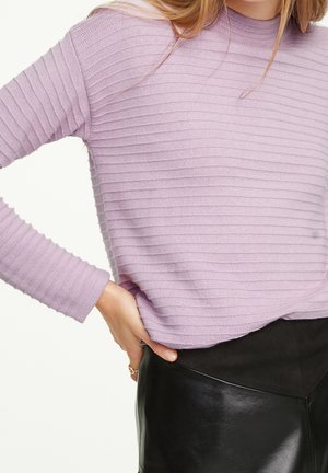 Kvinde iført en ribstrikket lavendelfarvet sweater med lange ærmer og et sort lædernederdel, hånden hvilende på hoften.