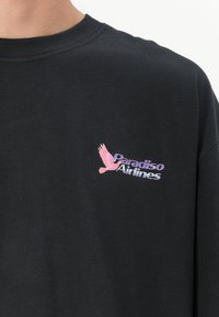 Zwarte katoenen t-shirt met een grafische print van een roze vogel en "Paradiso Airlines" in paars en wit. Korte mouwen en een ronde hals.