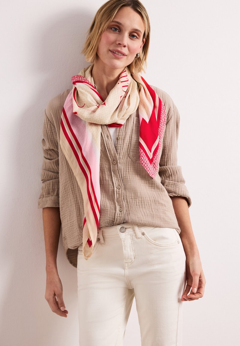 Femme portant une chemise beige à boutons, un pantalon blanc et une grande écharpe à motifs rouge, rose et beige drapée autour du cou.
