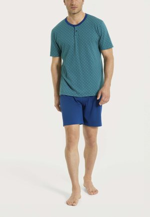 Uomo in piedi a piedi nudi che indossa una maglietta verde acqua a maniche corte con piccoli puntini e pantaloncini blu navy su uno sfondo semplice.