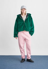 Grüne, flauschige Zip-Hoodie mit grauem Innenfutter, kombiniert mit weichen, rosa Jogginghosen und schwarzen Sneakers. Das Outfit hat einen lässigen Stil und eine entspannte Passform.