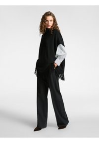 Zwarte poncho met kabelbrei-ontwerp, voorzien van franjes aan de zoom. Draagt over een gestreepte lange mouw shirt en gecombineerd met zwarte schoenen.
