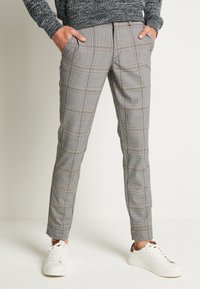 Pantalon à carreaux gris avec une coupe slim, présentant un motif à carreaux subtil dans des tons noirs et marron. Associé à des baskets blanches pour le contraste.