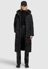 Zwarte lange pufferjas met een capuchon, voorzien van een rits- en drukknoopsluiting, zijzakken en een contrasterende logotape aan de voorkant. Gecombineerd met zwarte broek.