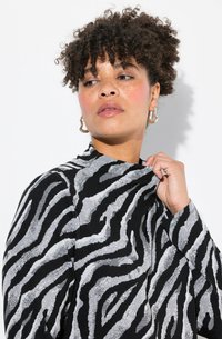 Zwart en zilver gestreepte zebra-jack met hoge kraag, gestructureerde stof, lange mouwen en een subtiele glans; model houdt de kraag vast.