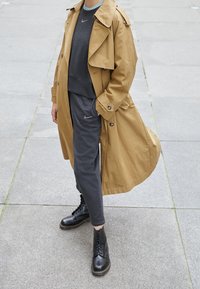 Beige trenchcoat över en mörkgrå sweatshirt och matchande byxor. Svarta snörboots med kraftig sula. Slät tyg, skräddarsydd passform.