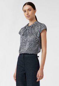 Graue Bluse mit Leopardenmuster, kurzen Ärmeln, gerafftem Ausschnitt und lockerer Passform. Kombiniert mit dunkelblauen, hochtaillierten Hosen.