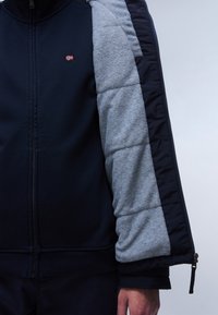 Granatowa kurtka na zamek z szarym rękawem z polaru. Posiada ściągacze przy mankietach oraz małe haftowane logo z przodu. Gładka faktura, nowoczesny design.