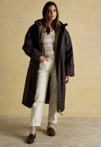 Sievietei mugurā ir garš tumši brūns mētelis, zem kura ir rakstaina džemperis, baltas bikses, bēšu zeķes un brūnas loafers, stāvot uz bēša fona.