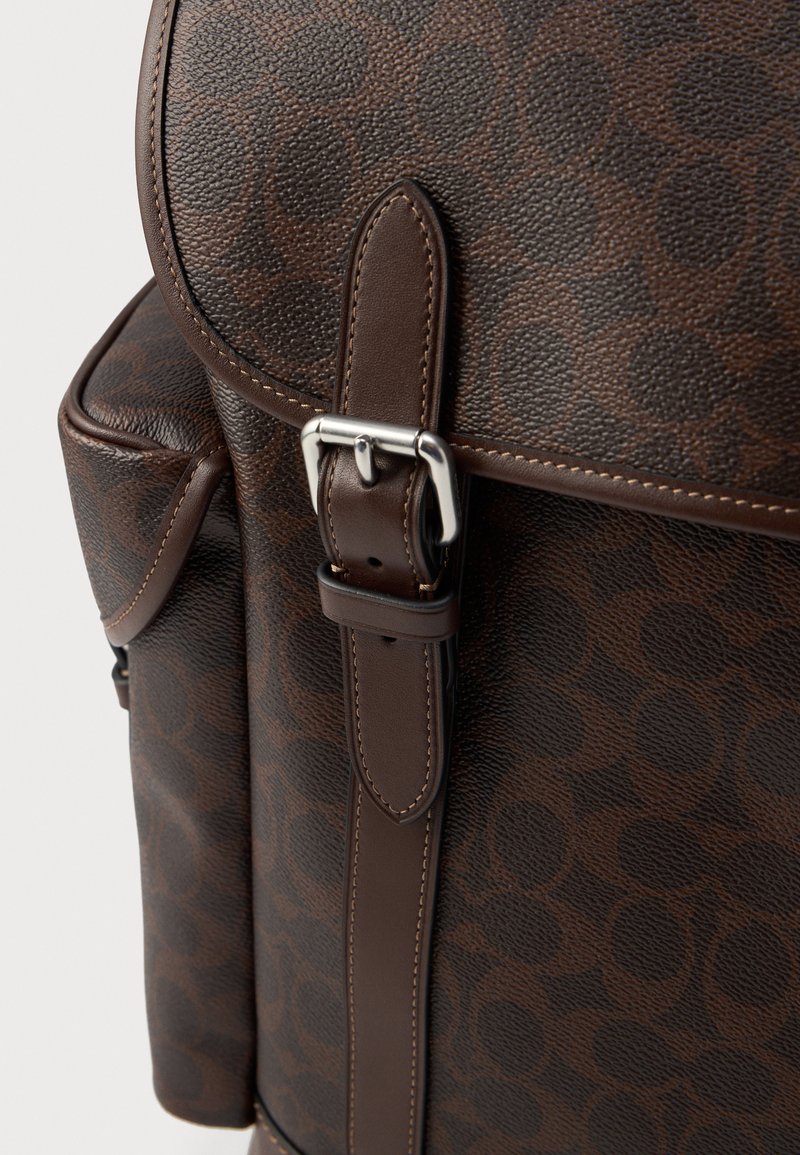 Brown leather backpack with a textured surface, featuring a circular pattern, silver hardware, and a strap with a metal buckle detail.