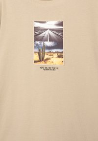 Beige bomulds T-shirt med et centralt grafisk motiv, der viser to billeder: en vej og en ørken scene med kaktusser, samt teksten "HER KAN DU FINDE MIG."