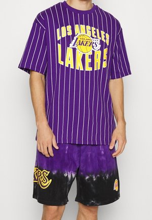 Une personne se tient debout portant un t-shirt rayé violet et blanc des LA Lakers avec des lettres jaunes et un short en tie-dye noir et violet.
