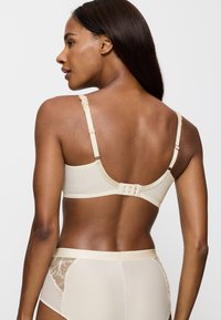 Beige rintaliivi, jossa on pitsikoristeet, säädettävät olkaimet ja kolme hakasta takana. Silkkinen pinta ja istuva muotoilu.