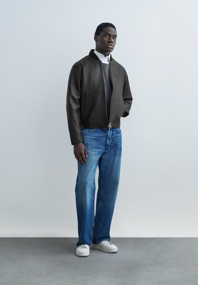 Άνδρας που στέκεται φορώντας μαύρο δερμάτινο μπουφάν πάνω από γκρι πουλόβερ και λευκό πουκάμισο, μπλε τζιν και λευκά αθλητικά παπούτσια, μπροστά σε ένα μονόχρωμο φόντο.