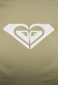 Συμμετρικό λευκό γεωμετρικό λογότυπο σε σχήμα καρδιάς με λεπτομέρειες που μοιάζουν με βέλη σε φόντο ελιάς πράσινου υφάσματος.