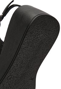 Semelle noire texturée d'une chaussure avec un talon plateforme épais et une boucle de bride de cheville visible sur un fond blanc.