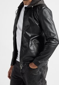 Černá kožená bunda s kapucí, opatřená zipem vpředu, bočními kapsami a hladkou texturou. Nositelná přes bílé tričko a šedé džíny.