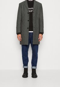 Abrigo de espiga gris, abotonado, usado sobre un suéter negro con logo. Combinado con jeans azules remangados y botas negras de cordones.