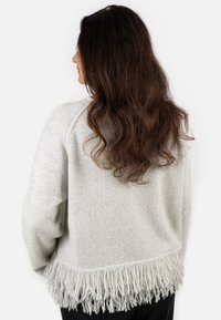 Frau mit langen, welligen braunen Haaren, die einen hellgrauen Pullover mit Fransen am Saum trägt, von hinten und in Seitenansicht gezeigt.