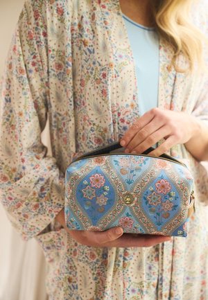 Persoon met een bloemenpatroon badjas opent een blauwe cosmetische tas met bloemenprint, voorzien van een gouden rits en centraal embleem.