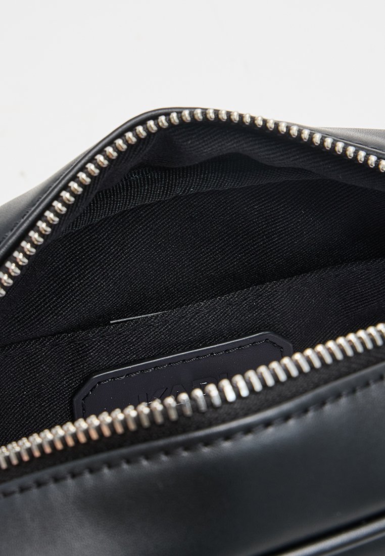 Bolso de cuero negro con una cremallera plateada. El interior cuenta con un forro de tela negra lisa y una etiqueta. Diseño sencillo sin patrones visibles.